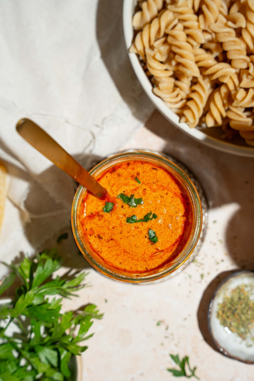 A jar of creamy tomato pasta sauce garnished with fresh basil. There is a spoon in the jar. The jar is on a tan counter with a bowl of fusilli pasta and white cloth napkin.