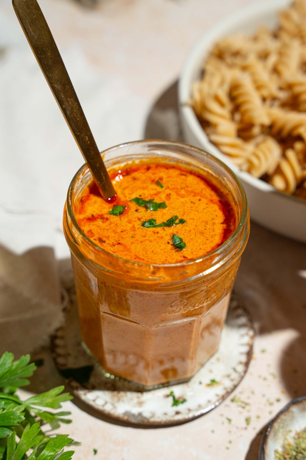 A jar of creamy tomato pasta sauce garnished with fresh basil. There is a spoon in the jar. The jar is on a tan counter with a bowl of fusilli pasta and white cloth napkin.