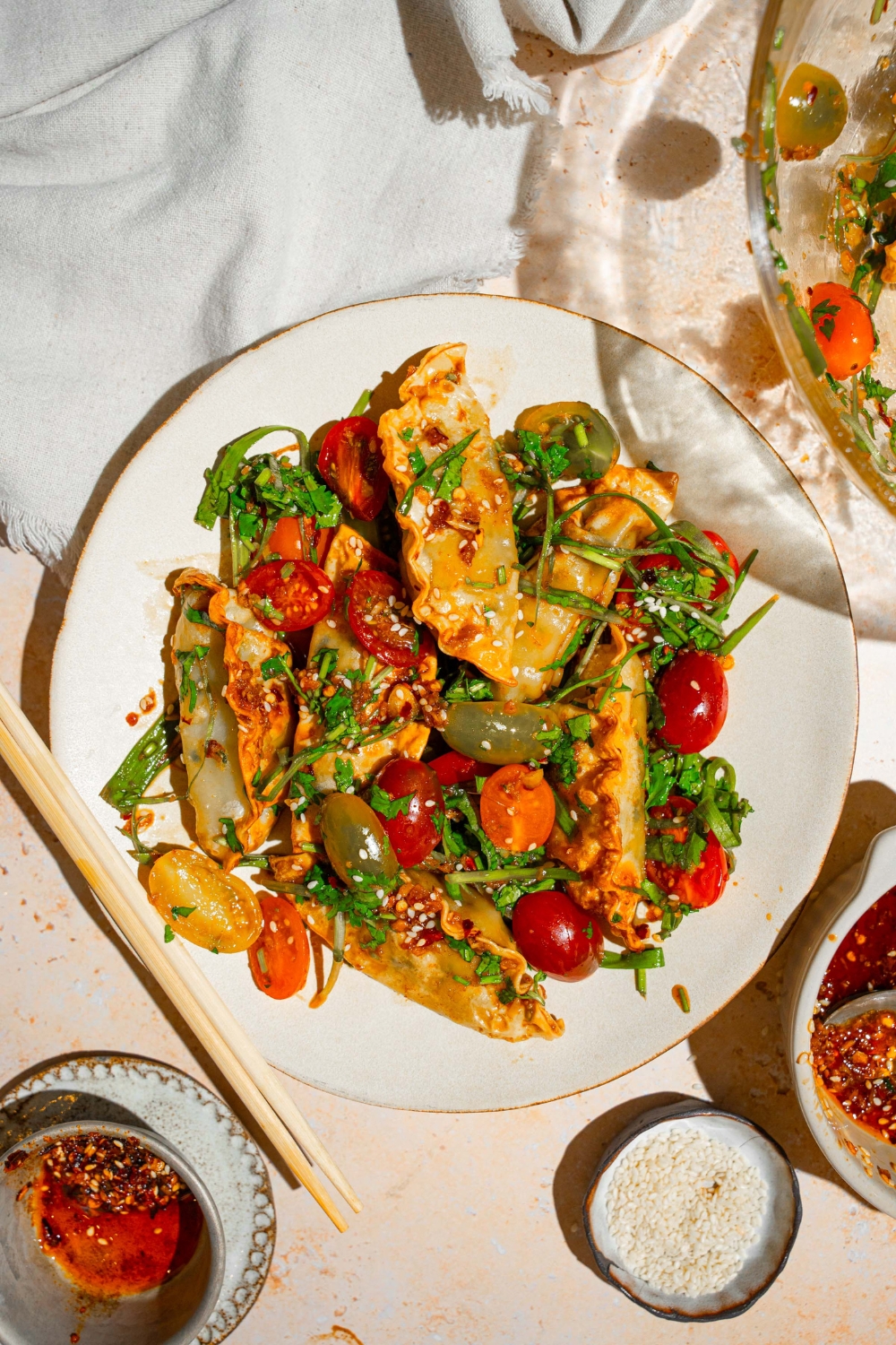 A white plate with crispy dumpling salad with crispy dumplings, sliced tomatoes, and scallions, tossed in a soy sauce and chili oil dressing. The plate is on a tan counter with a pair of chopsticks and a white cloth napkin.