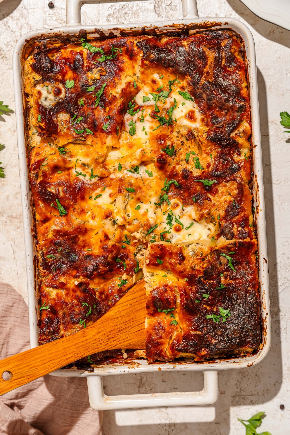 A baking dish with baked chicken alfredo lasagna garnished with fresh parsley. A wooden spoon is removing a slice from the dish. The dish is on a tan counter with a brown cloth napkin.