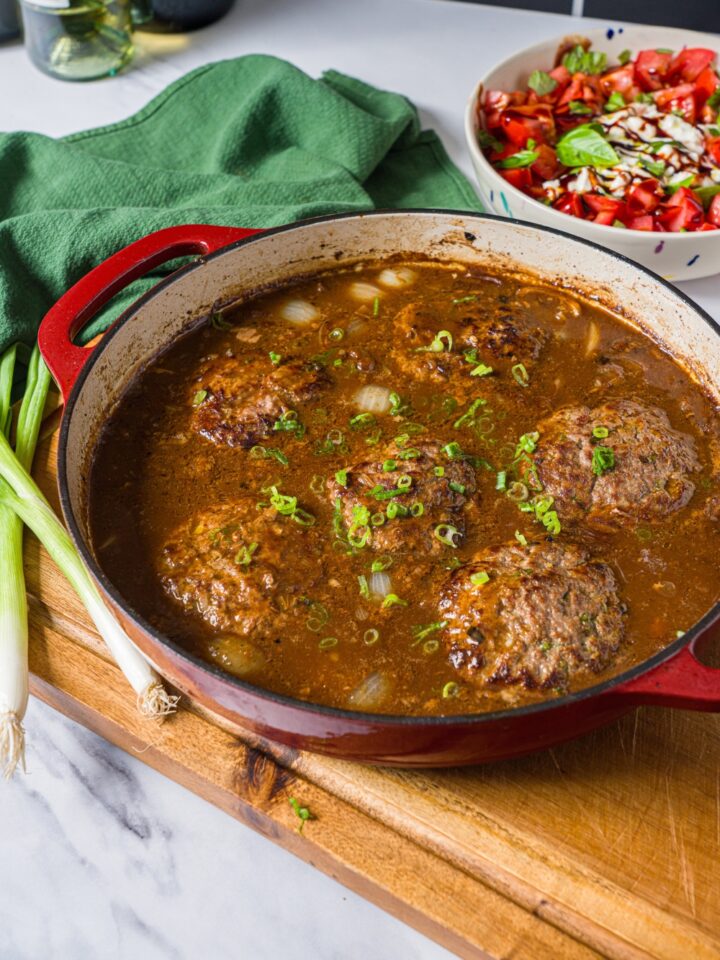A cast iron skillet with hamburger patties in onion gravy garnished with sliced green onion. The skillet is on a wooden board with a green cloth napkin.