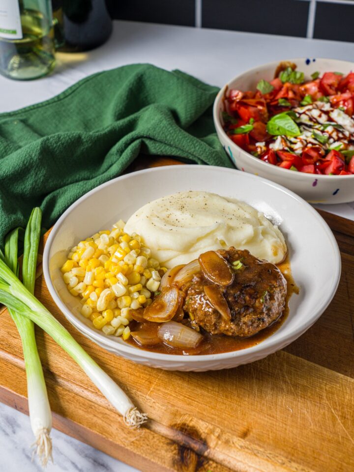 A white plate with hamburger steak in onion gravy served with mashed potatoes and corn. The plate is on a wooden board with a green cloth napkin.