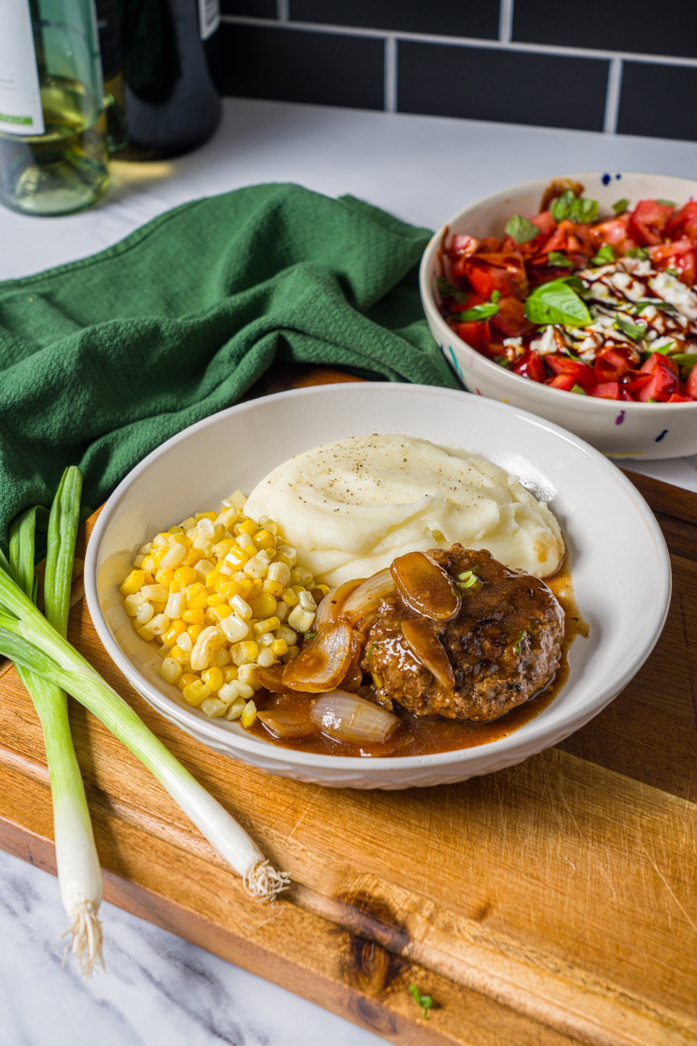 A white plate with hamburger steak in onion gravy served with mashed potatoes and corn. The plate is on a wooden board with a green cloth napkin.