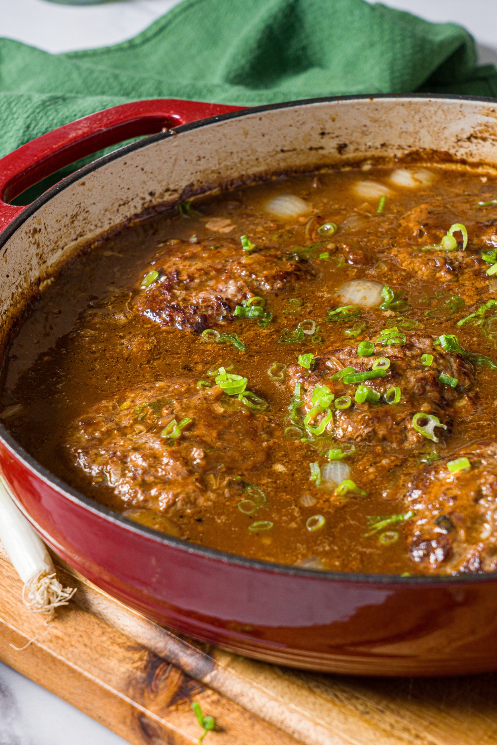 A cast iron skillet with hamburger patties in onion gravy garnished with sliced green onion. The skillet is on a wooden board with a green cloth napkin.