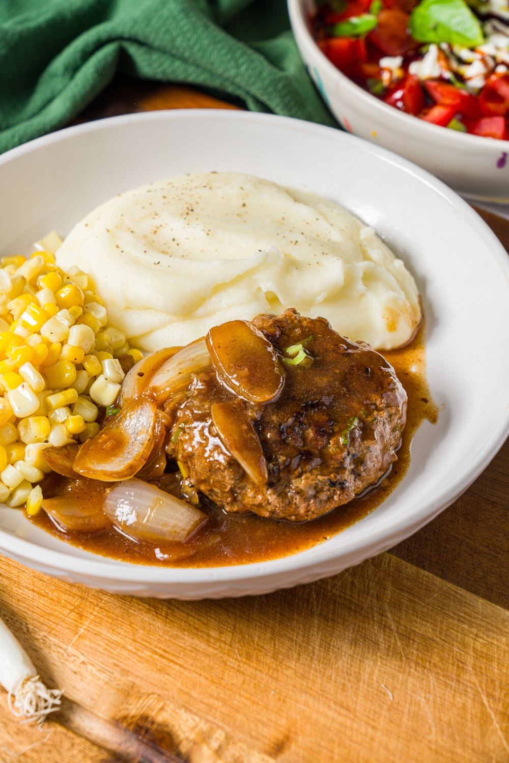 A white plate with hamburger steak in onion gravy served with mashed potatoes and corn. The plate is on a wooden board with a green cloth napkin.