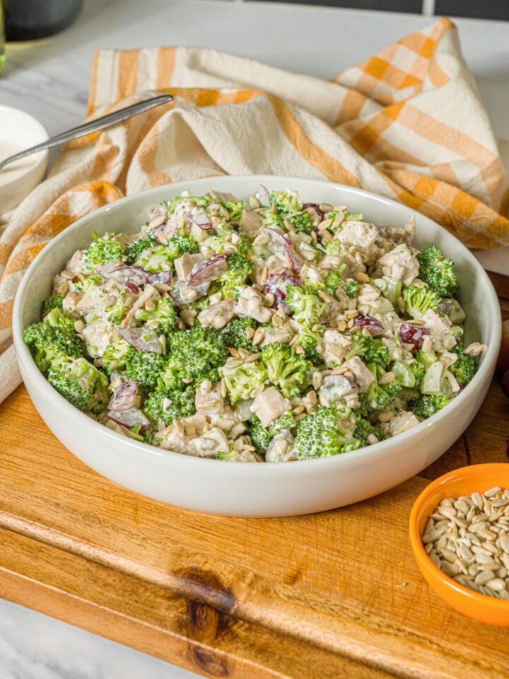 A healthy high protein broccoli chicken salad in a white bowl on top of a wooden cutting board.