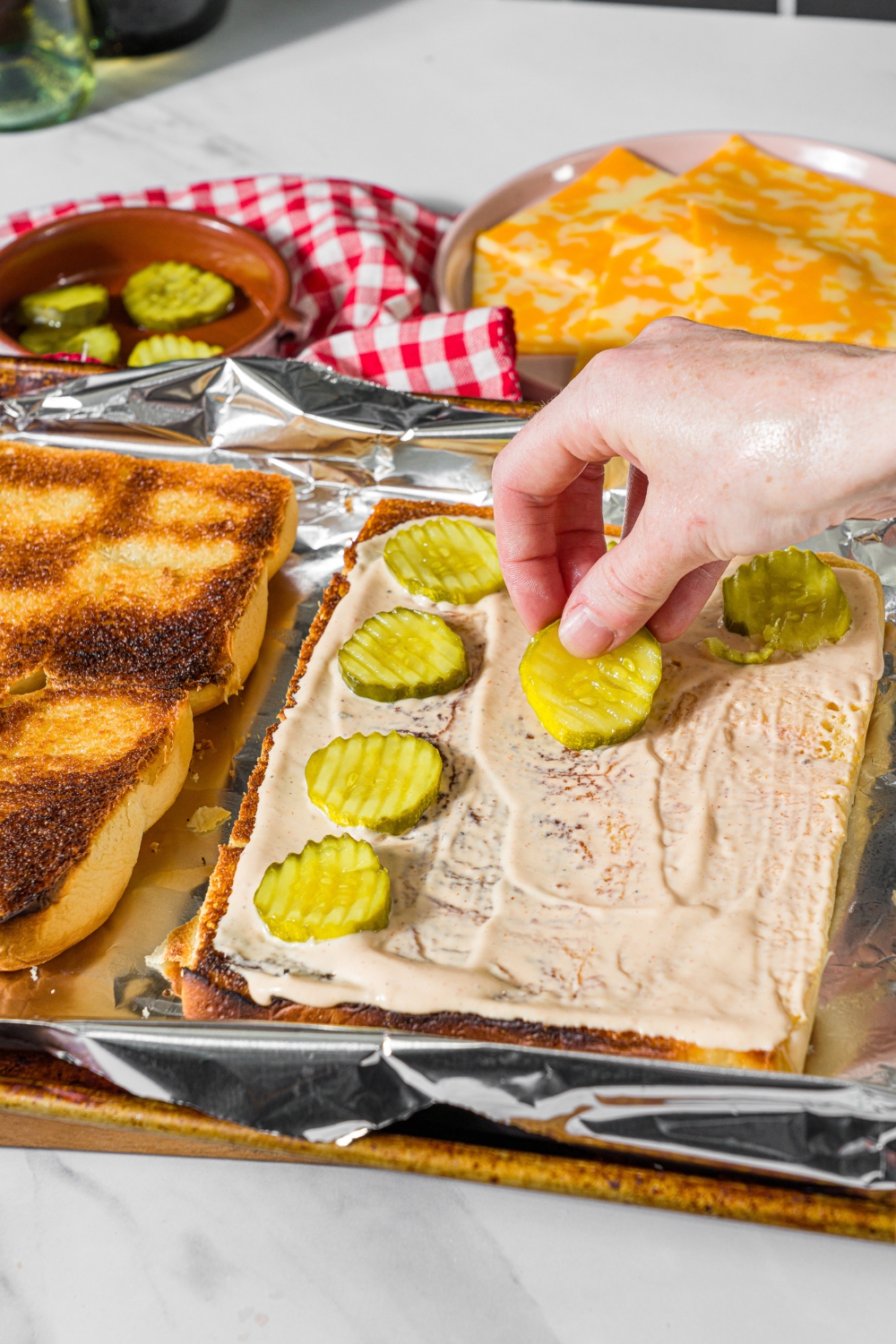 A baking sheet lined with foil with sliced toasted hawaiian rolls. The bottom rolls are spread with burger sauce with pickles being added on top.