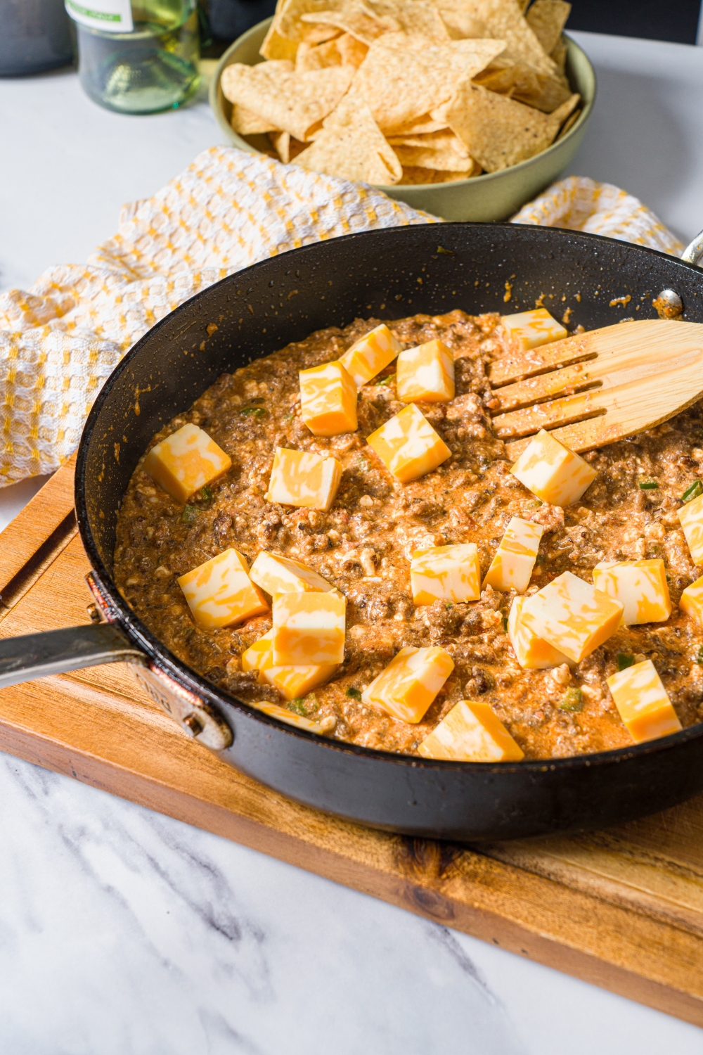 A skillet with seasoned ground beef mixture topped with cheese cubes. The skillet is on a wooden board on a marble counter with a bowl of chips.
