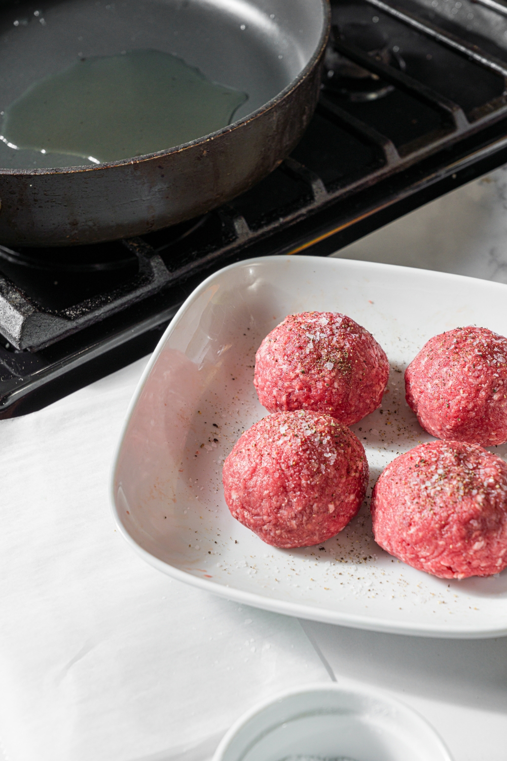 A skillet heating oil on a stove next to a marble counter with a bowl of seasoned ground beef patty balls.