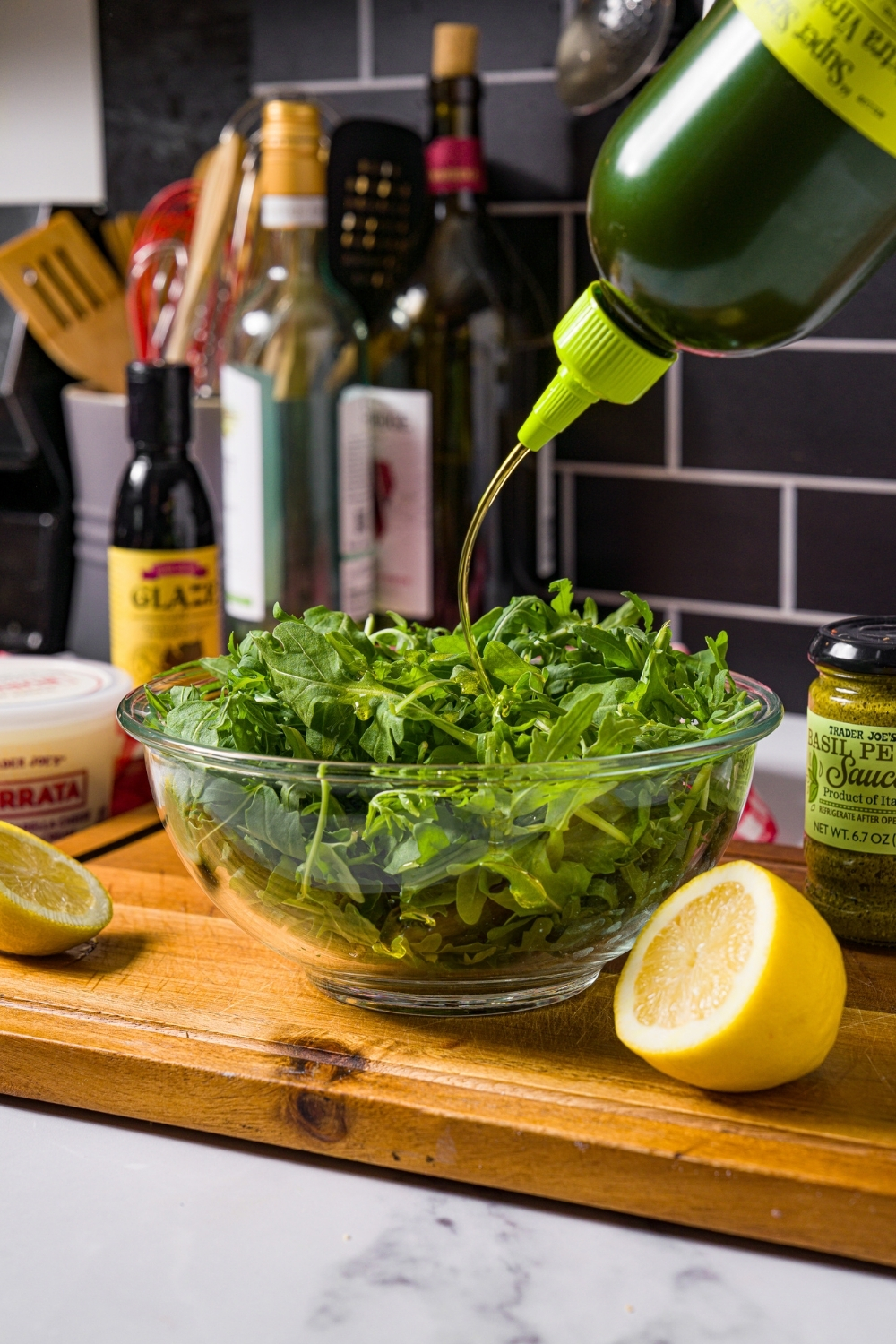 A bottle of olive oil being poured into a glass bowl that is filled with arugula.