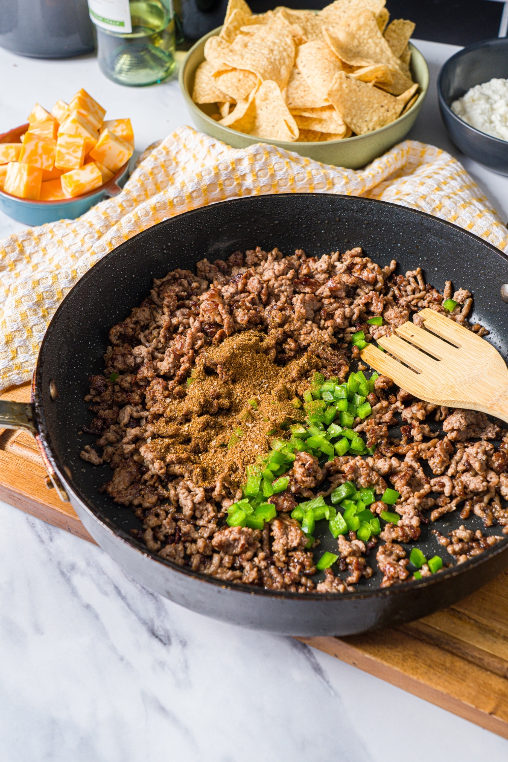 A skillet with a slotted spoon mixing ground beef with seasonings and chopped jalapeño. The skillet is on a wooden board on a marble counter with a bowl of chips.