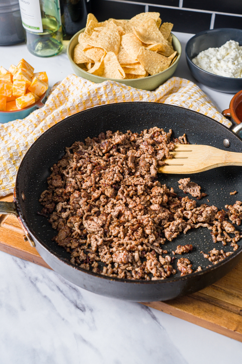 A skillet with a slotted spoon mixing ground beef. The skillet is on a wooden board on a marble counter with a bowl of chips.