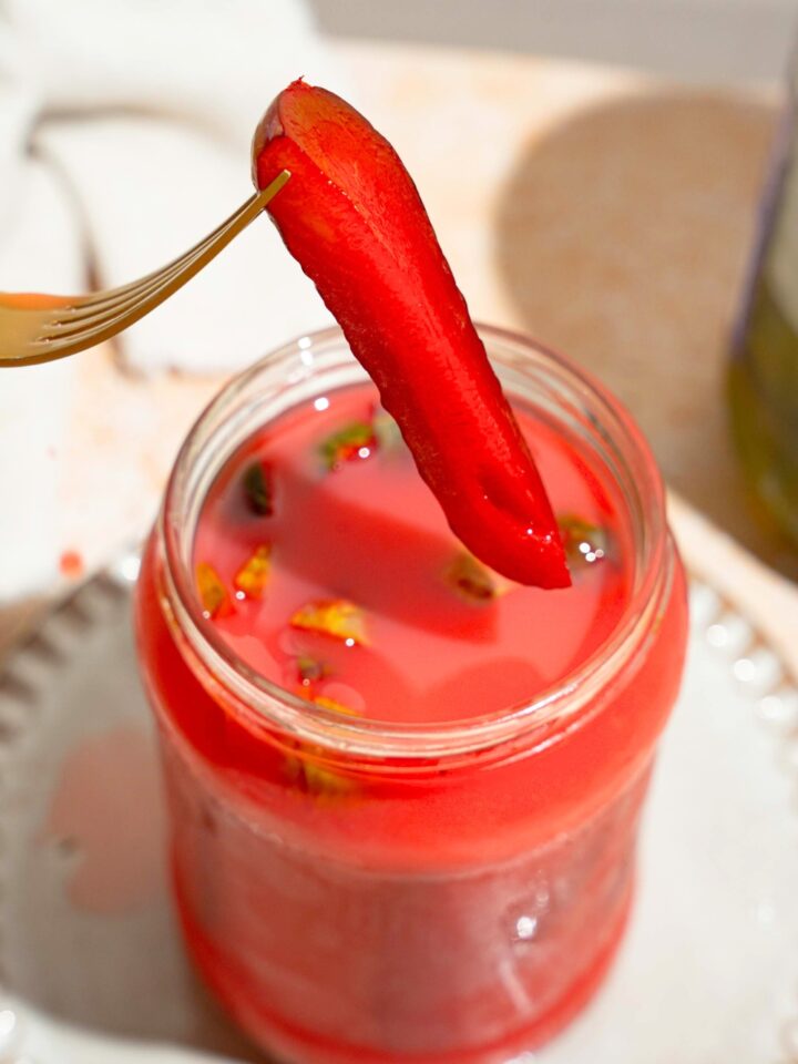 A kool aid pickle spear with a fork in it being held above a jar that is filled with the koolickles.