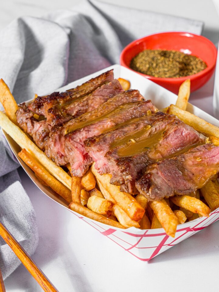 A fry basket with french fries topped with sliced steak and topped with steak frites sauce. The basket is on a marble counter with a bowl of seasonings and a gray cloth napkin.