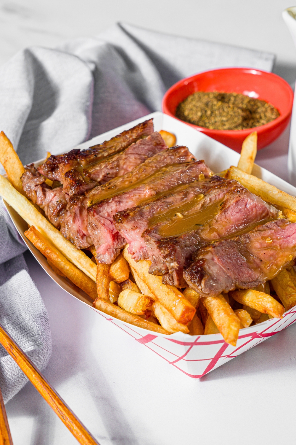 A fry basket with french fries topped with sliced steak and topped with steak frites sauce. The basket is on a marble counter with a bowl of seasonings and a gray cloth napkin.