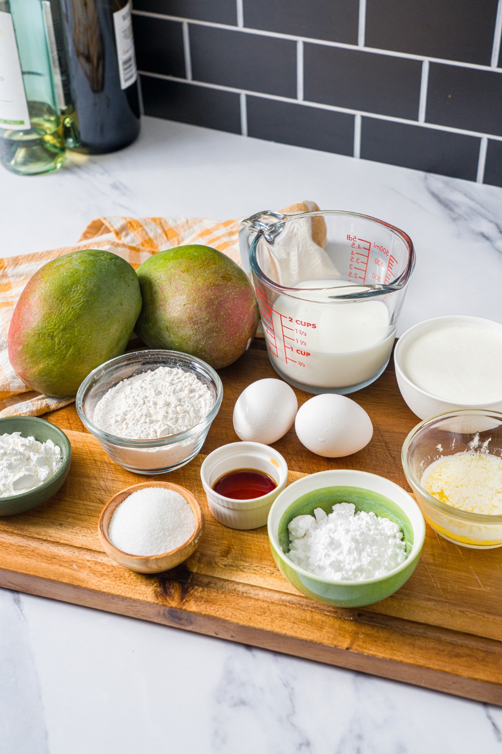 Two mangos, a pitcher of heavy cream, a bowl of powdered sugar, a bowl of flour, two eggs, vanilla extract, and sugar all on a wood cutting board on a white counter.