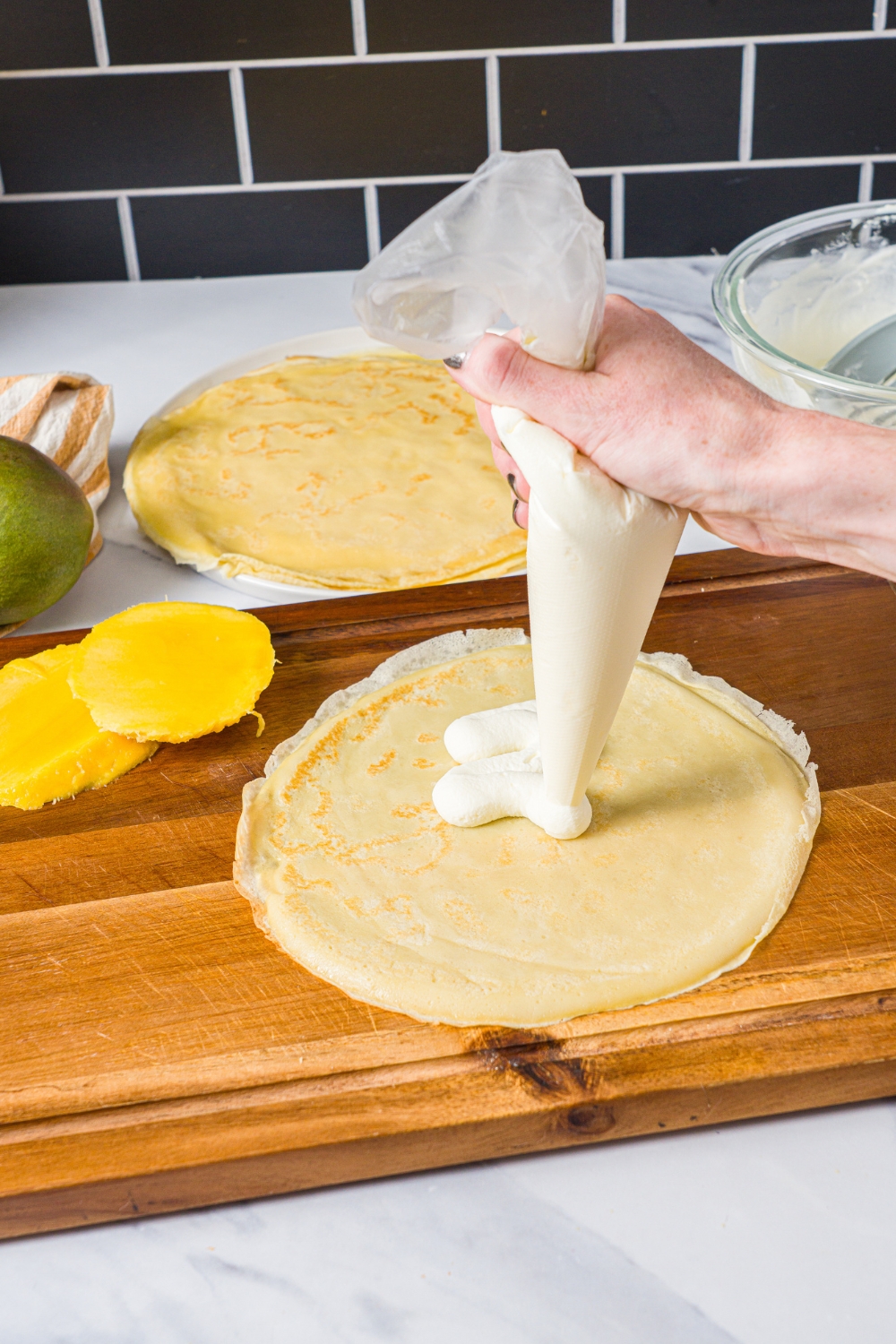 A hand piping a cream mixture onto a crêpe for mango pancakes.
