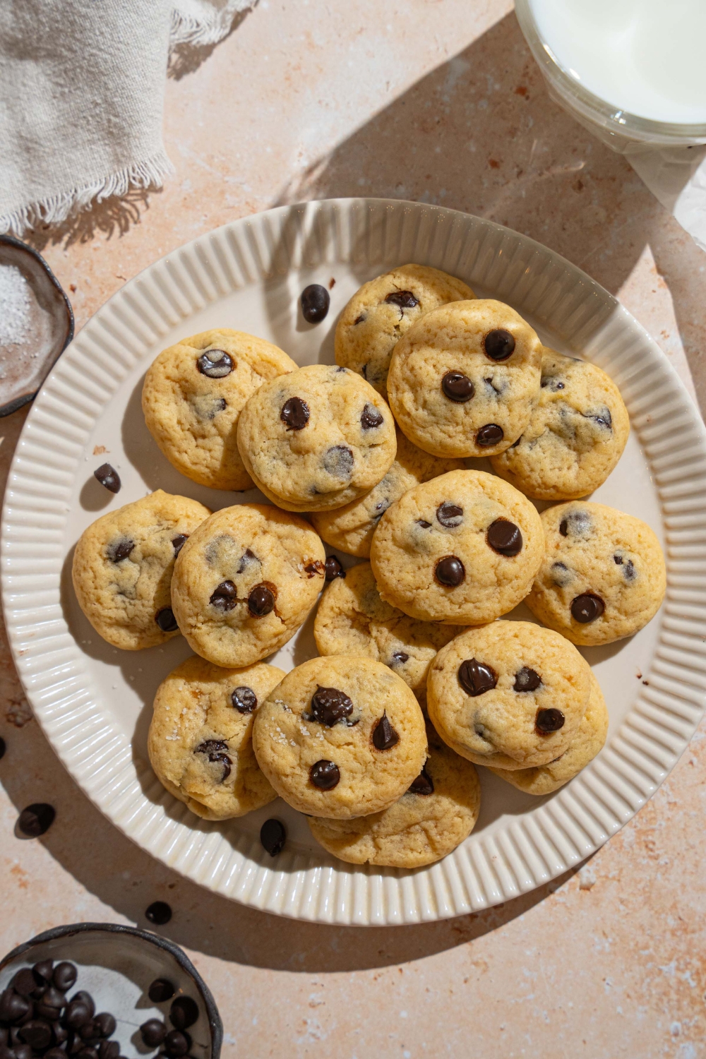 A plate of mini chocolate chip cookies on a tan counter with a glass of milk and bowl of chocolate chips.