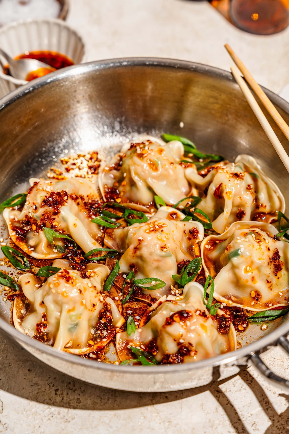 A skillet with pan fried pork and shrimp dumplings topped with sliced green onion, sesame seeds, and a soy sauce chili oil mixture. The skillet is on a tan counter with a pair of chopsticks and a plate of garnishes.
