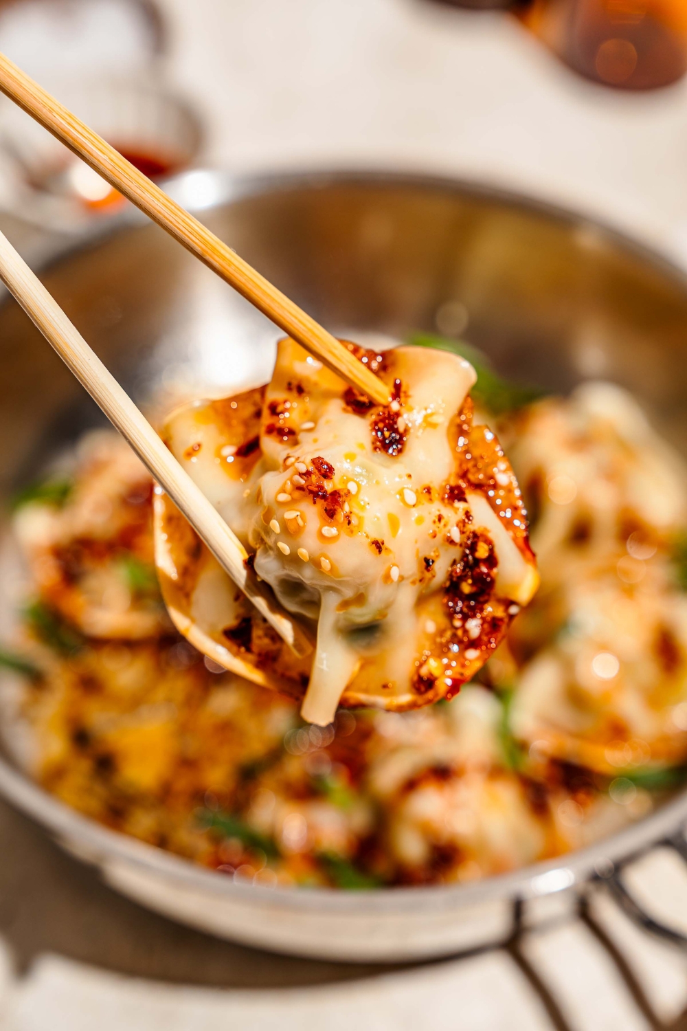 A close up of a pair of chopsticks holding a pan fried pork and shrimp dumpling garnished with sesame seeds and served with a soy sauce and chili oil mixture. There is a skillet of dumplings blurred in the background.