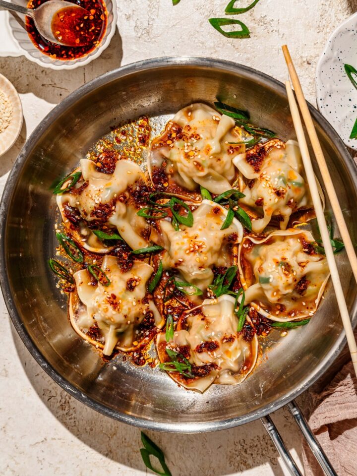 A skillet with pan fried pork and shrimp dumplings topped with sliced green onion, sesame seeds, and a soy sauce chili oil mixture. The skillet is on a tan counter with a pair of chopsticks and a plate of garnishes.
