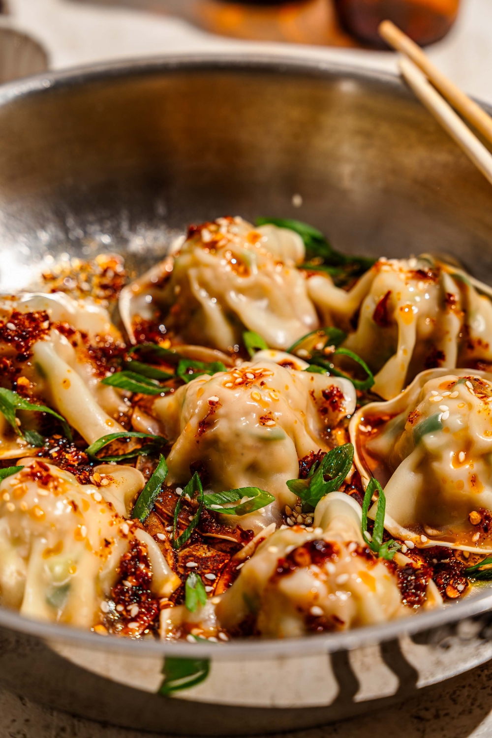 A skillet with pan fried pork and shrimp dumplings topped with sliced green onion, sesame seeds, and a soy sauce chili oil mixture. The skillet is on a tan counter with a pair of chopsticks.