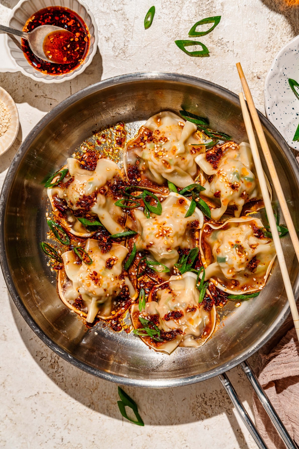 A skillet with pan fried pork and shrimp dumplings topped with sliced green onion, sesame seeds, and a soy sauce chili oil mixture. The skillet is on a tan counter with a pair of chopsticks and a plate of garnishes.