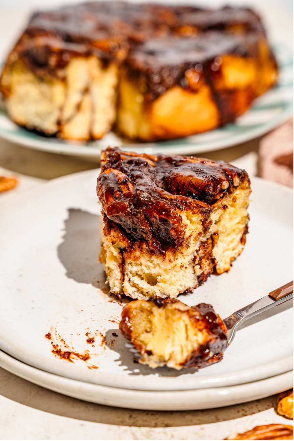 A close up of a white plate with a pecan caramel sticky bun with a fork taking a bite. There is a tray of sticky buns blurred in the background.