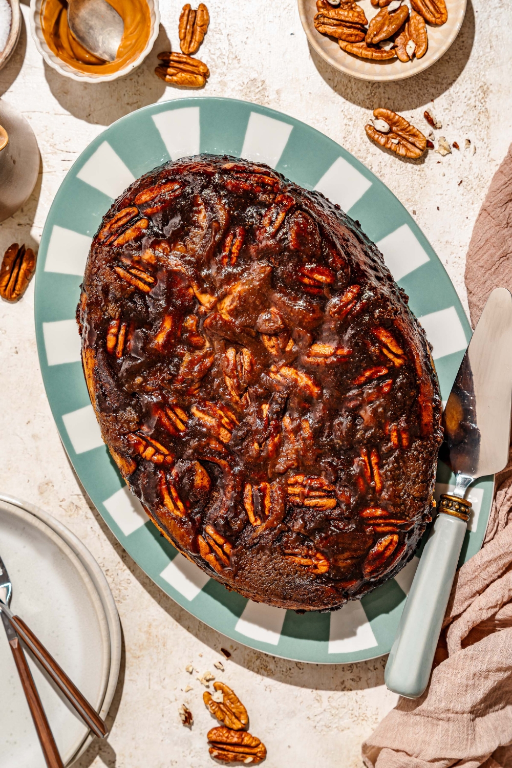 An oval tray with pecan caramel sticky buns. A bun has been removed from the tray. The tray is on a tan counter with bowls of pecans and cinnamon.