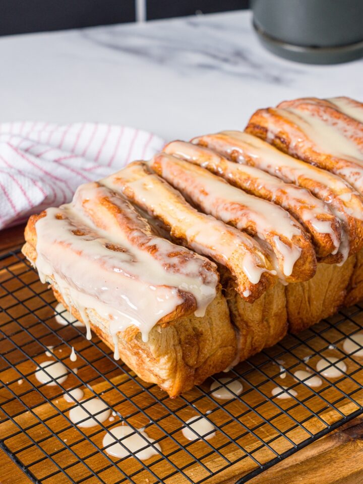 A loaf of pull apart cinnamon bread on a wire rack on top of a board with maple glaze on top and dripping off onto the wooden board.
