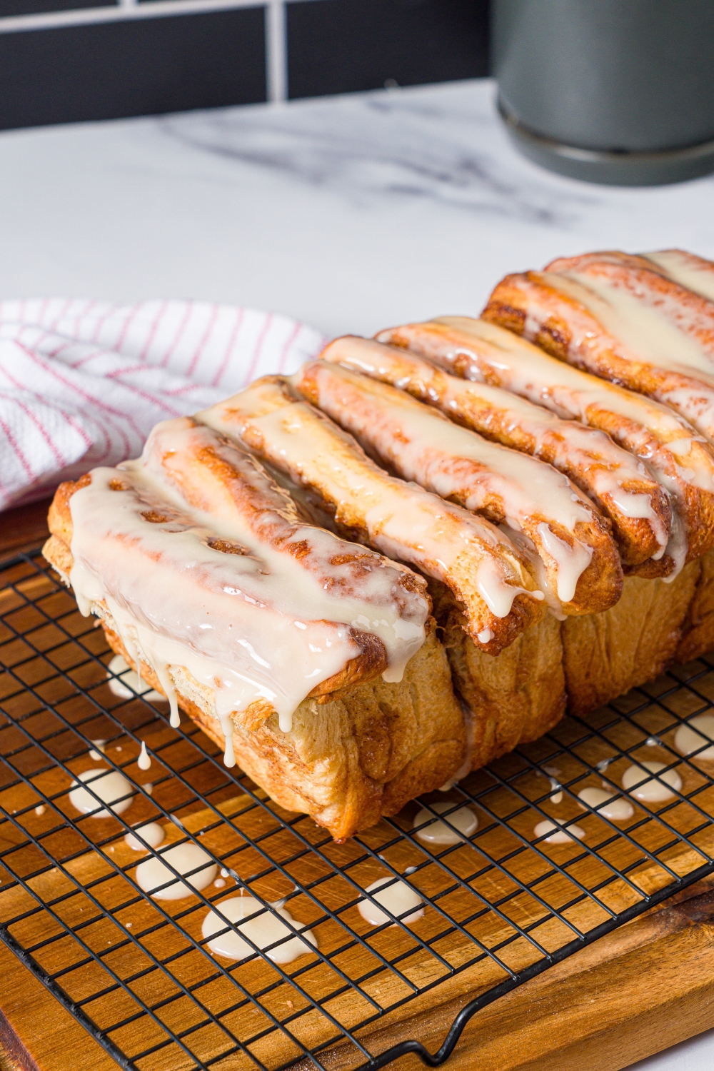 A loaf of pull apart cinnamon bread on a wire rack on top of a board with maple glaze on top and dripping off onto the wooden board.