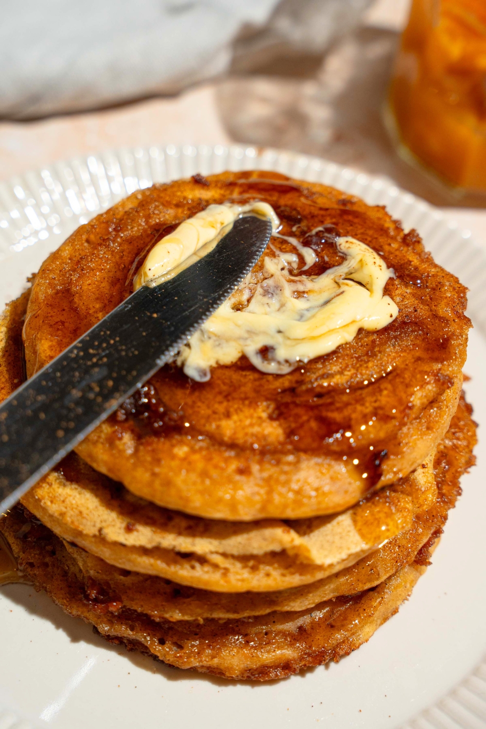 A knife spreading butter on a pumpkin cinnamon roll pancake that is on top of a few more pumpkin pancakes on a white plate.