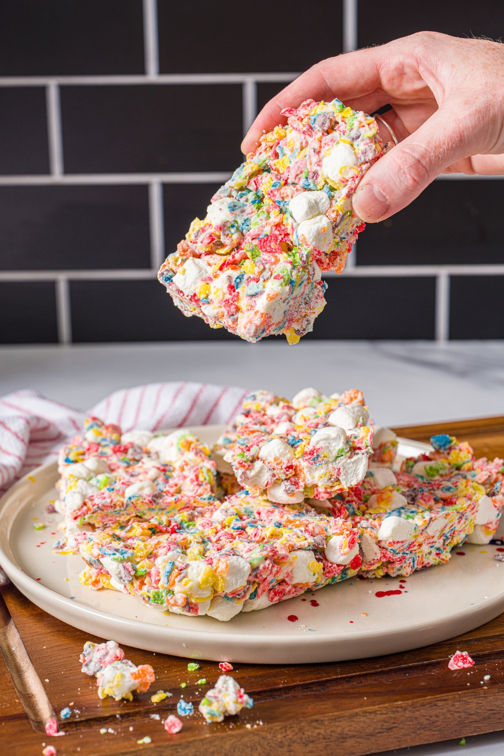 A white plate with fruity pebbles rice crispy treats. A hand is taking a treat from the plate. The plate is on a wooden board with a red striped napkin.