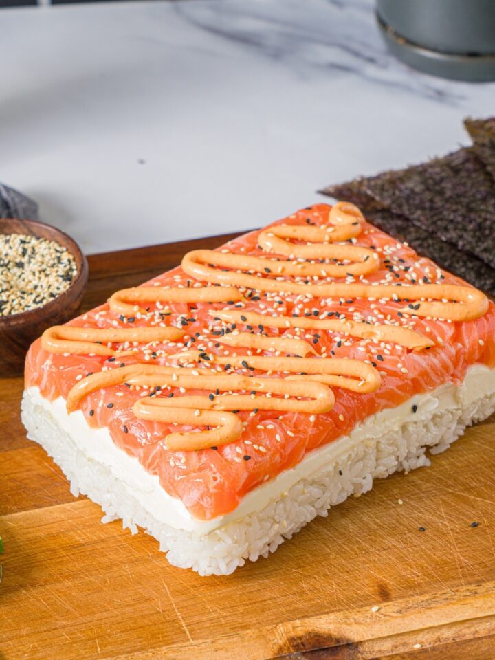 A salmon sushi cake with spicy mayo drizzled on top of salmon layered on cream cheese and rice on top of a wooden board.