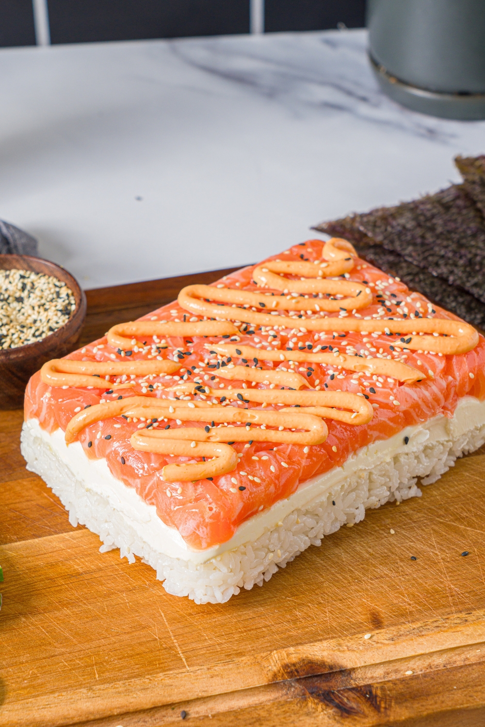 A salmon sushi cake with spicy mayo drizzled on top of salmon layered on cream cheese and rice on top of a wooden board.