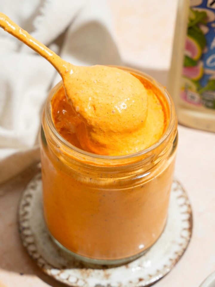 A spoon that has southwest chipotle sauce on it being held directly above a glass jar that is filled with the southwest chipotle sauce.