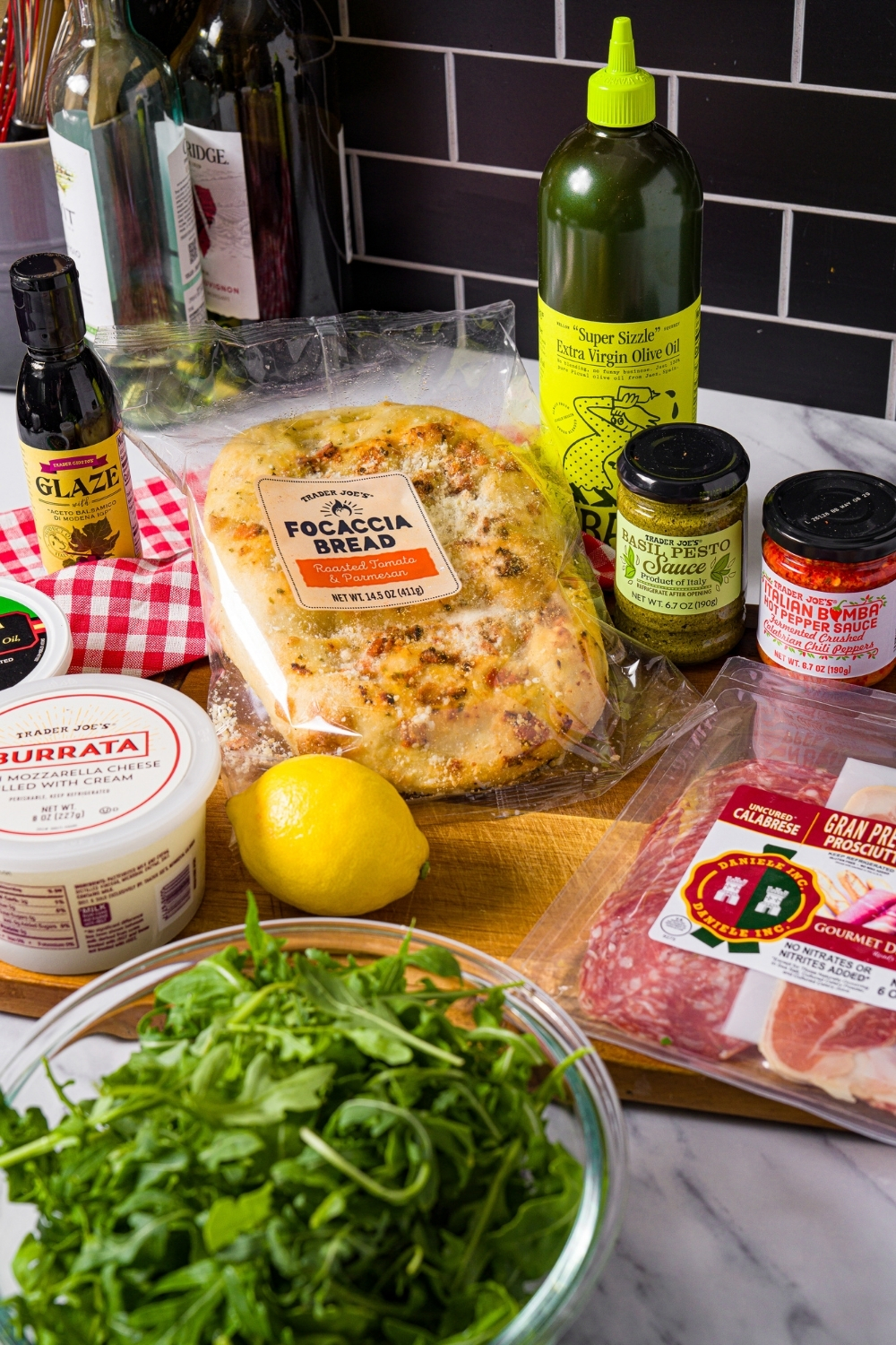 A package of Trader Joe's, focaccia, a pack of Trader Joe's, Barada, Trader Joe's italian, hot pepper sauce, Trader Joe's, basil, pesto sauce, a bowl of arugula, and a lemon on a cutting board.