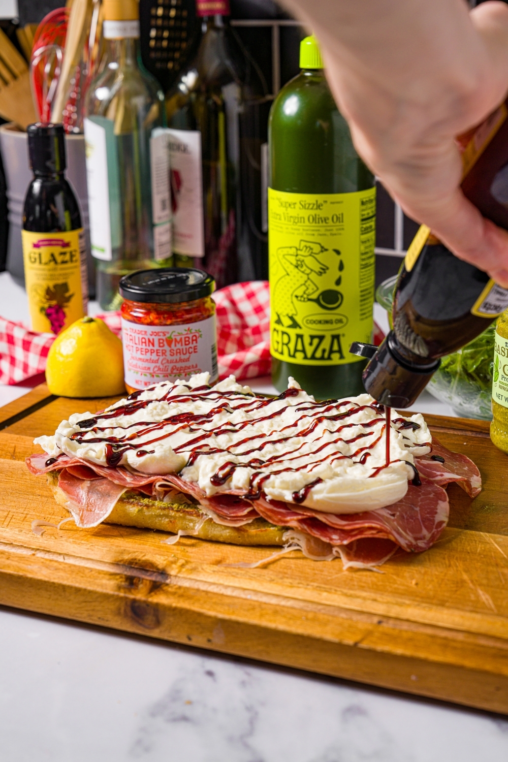 A hand holding a bottle of balsamic drizzle it on top of buratta that is deli meat layered on a slice of focaccia.
