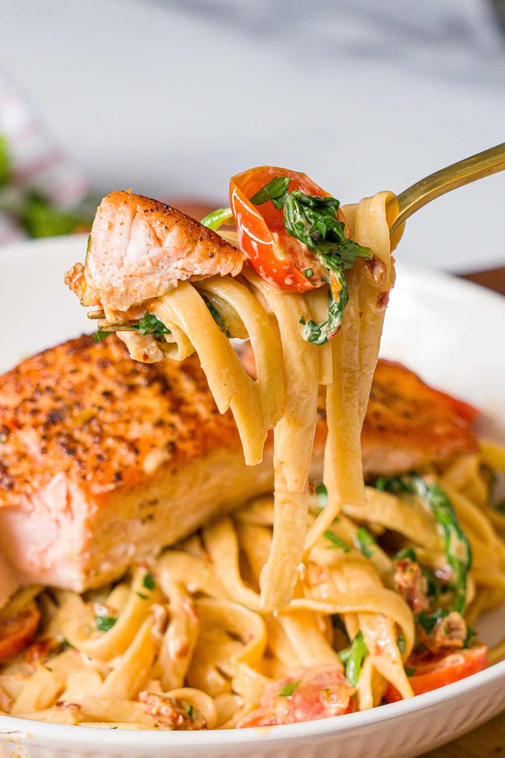A close up of a fork pulling a bite of creamy tuscan salmon with pasta. There is a bowl of salmon pasta on a wooden board blurred in the background.