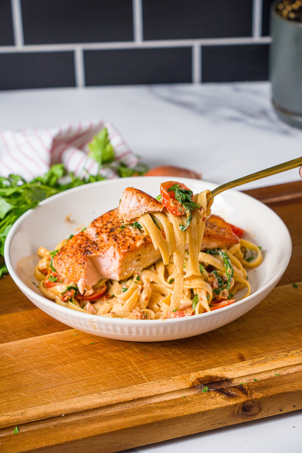 A white bowl with tuscan salmon with pasta. A fork is pulling a bite of pasta from the bowl. The bowl is on a wooden board.