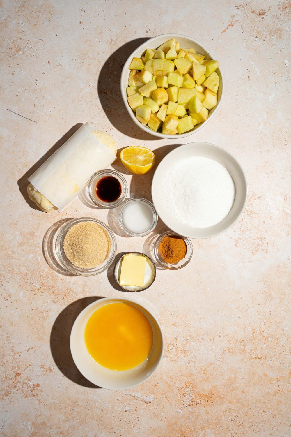 A tan counter with several bowls containing ingredients to make apple pie pull apart bread including apples, sugar, frozen biscuits, butter, lemon, cinnamon, and flour.