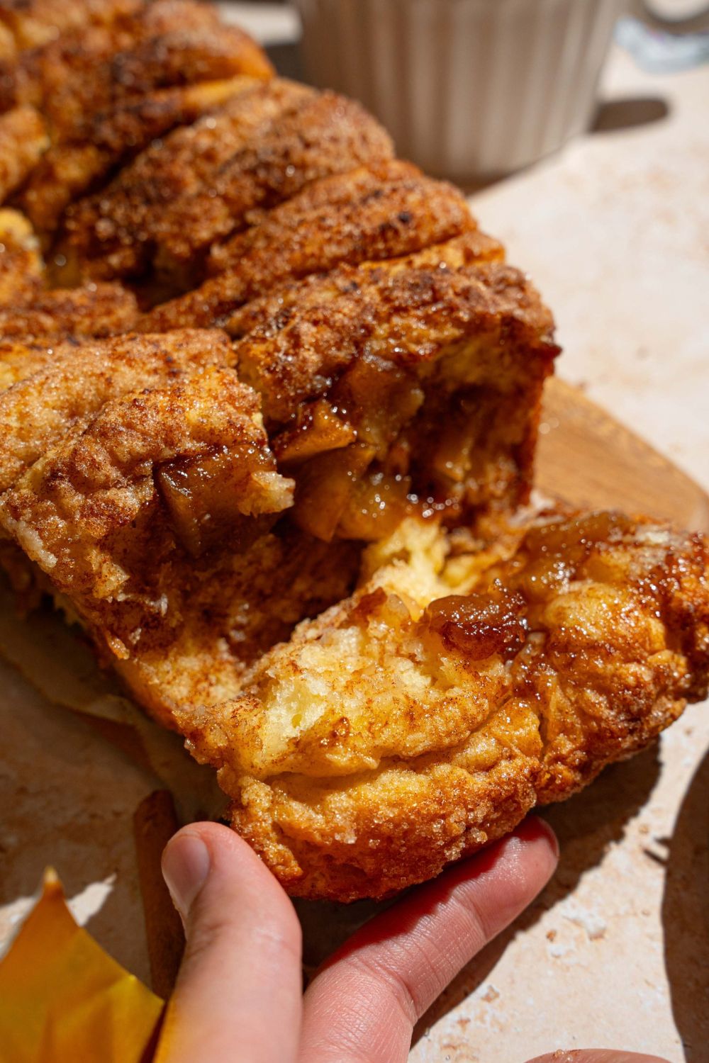 A loaf of apple pie pull apart bread on a wooden board lined with parchment paper. A hand is pulling a slice from the loaf. The board is on a tan counter.