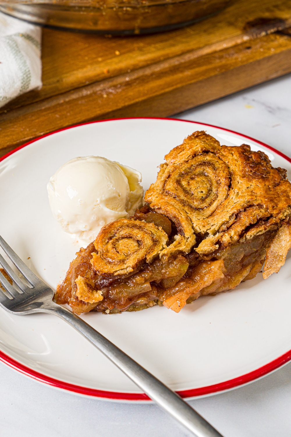 A white plate with a slice of apple pie with cinnamon roll crust served with a scoop of vanilla ice cream and a fork. The plate is on a marble counter.