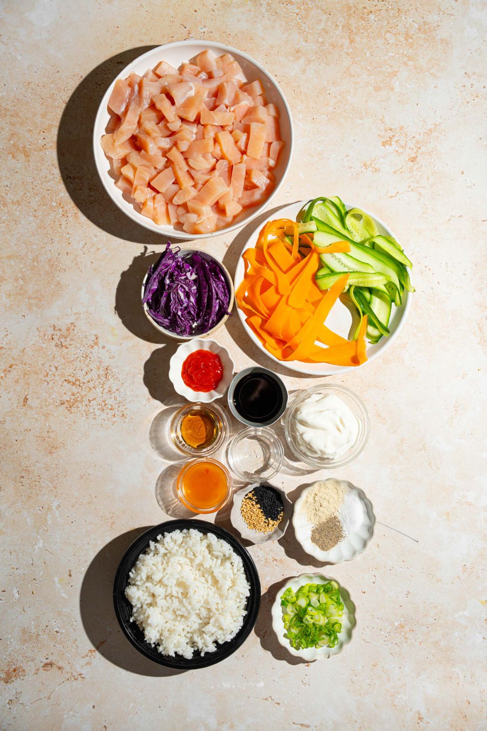 A tan counter with bowls containing ingredients to make bang bang chicken bowls including chicken, rice, cabbage, carrots, cucumber, green onion, mayo, thai chili sauce, sriracha, honey, and more.