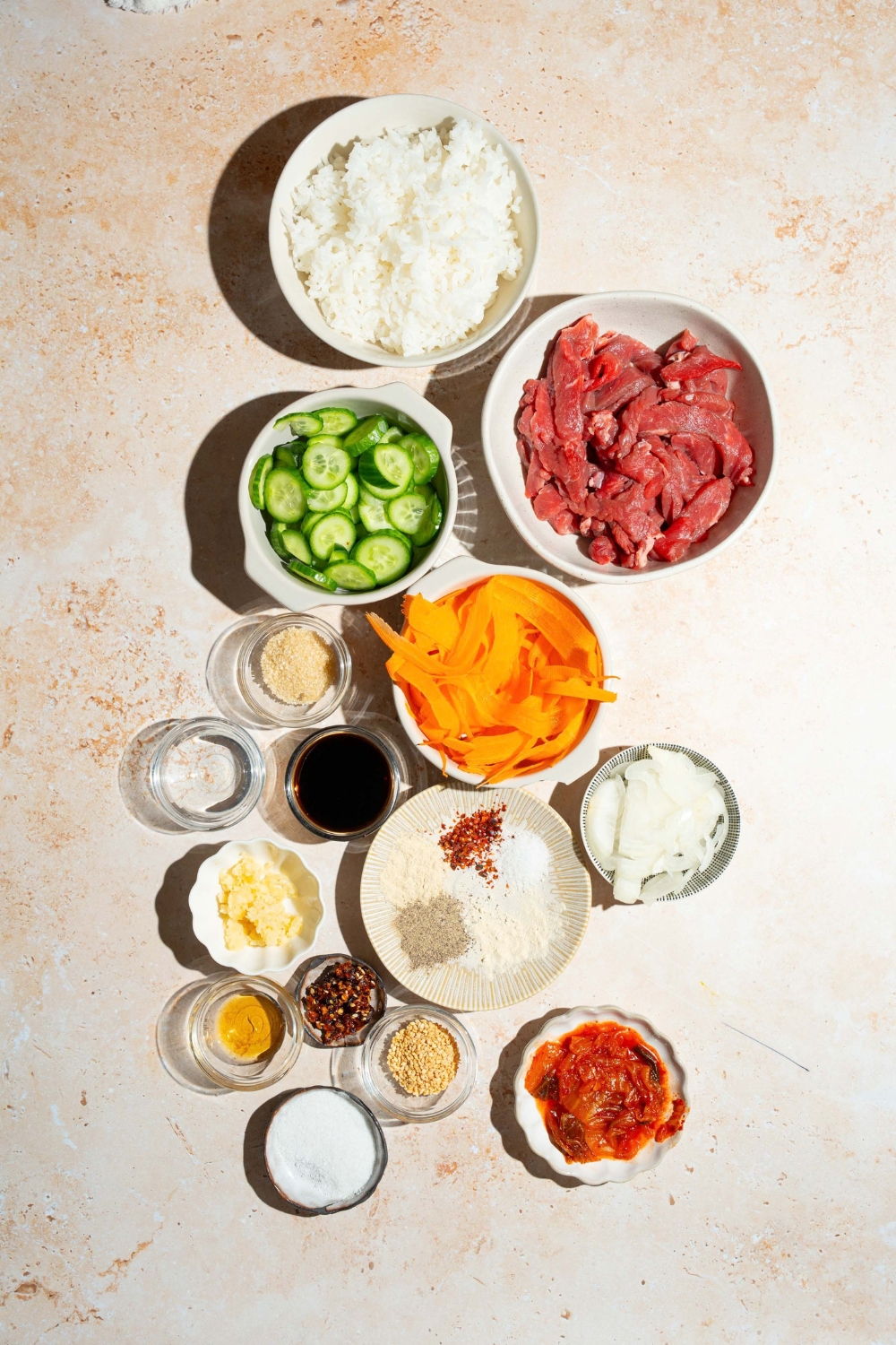 A tan counter with several bowls of ingredients to make beef bulgogi bowls including beef, cucumbers, carrots, rice, soy sauce, kimchi, rice vinegar, onions, and seasonings.