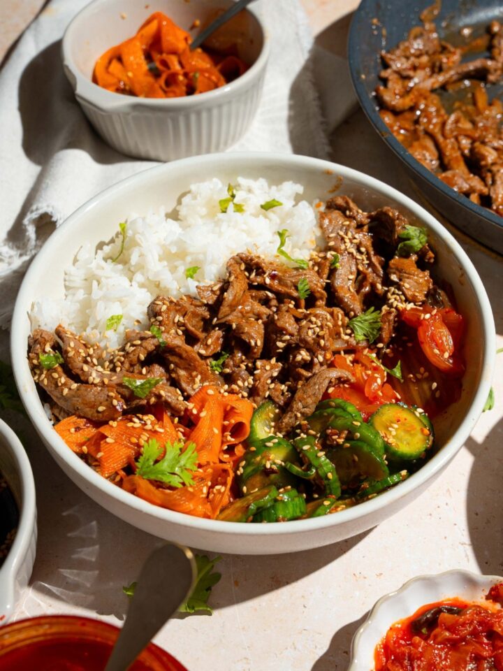 A white bowl with a beef bulgogi bowl served with white rice, beef bulgogi, and a cucumber carrot salad, all garnished with sesame seeds. The bowl is on a tan counter with bowls of ingredients.