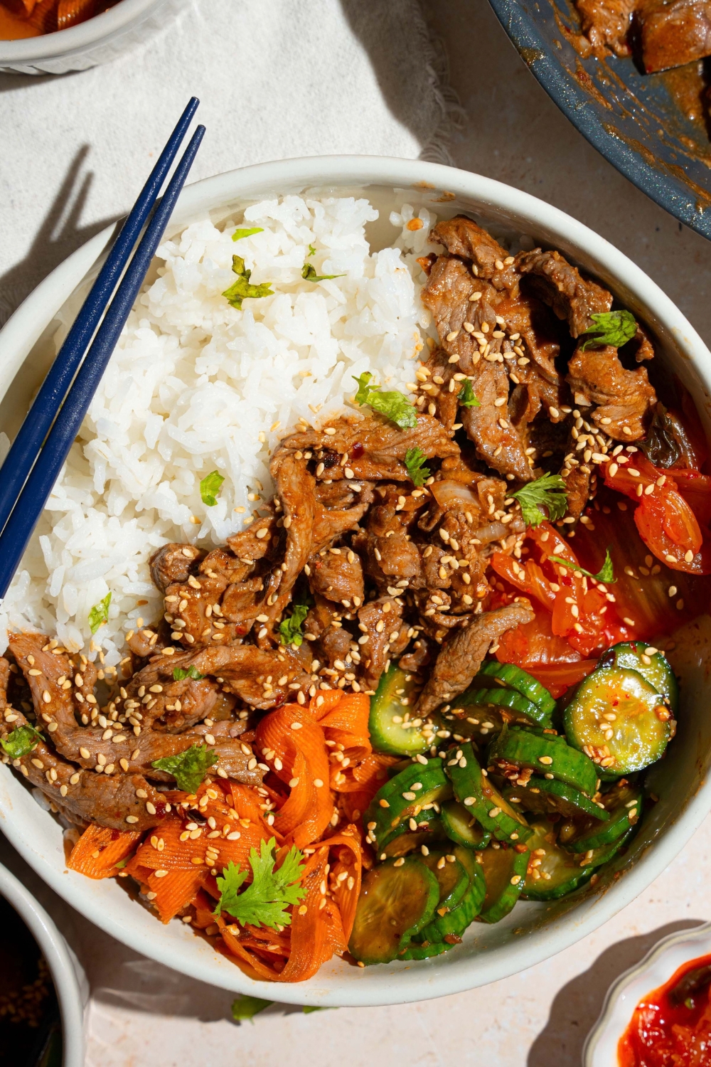 A white bowl with a beef bulgogi bowl made with rice, beef bulgogi, and a cucumber carrot salad, garnished with sesame seeds. A pair of chopsticks are on the bowl. The bowl is on a tan counter.