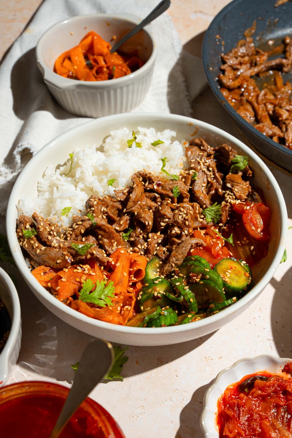 A white bowl with a beef bulgogi bowl served with white rice, beef bulgogi, and a cucumber carrot salad, all garnished with sesame seeds. The bowl is on a tan counter with bowls of ingredients.