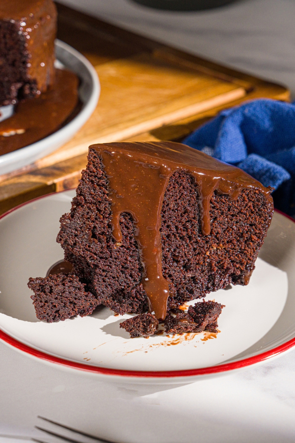 A white plate with a slice of Matilda cake topped with chocolate ganache. The plate is on a marble counter with a wooden board with a plate of cake.