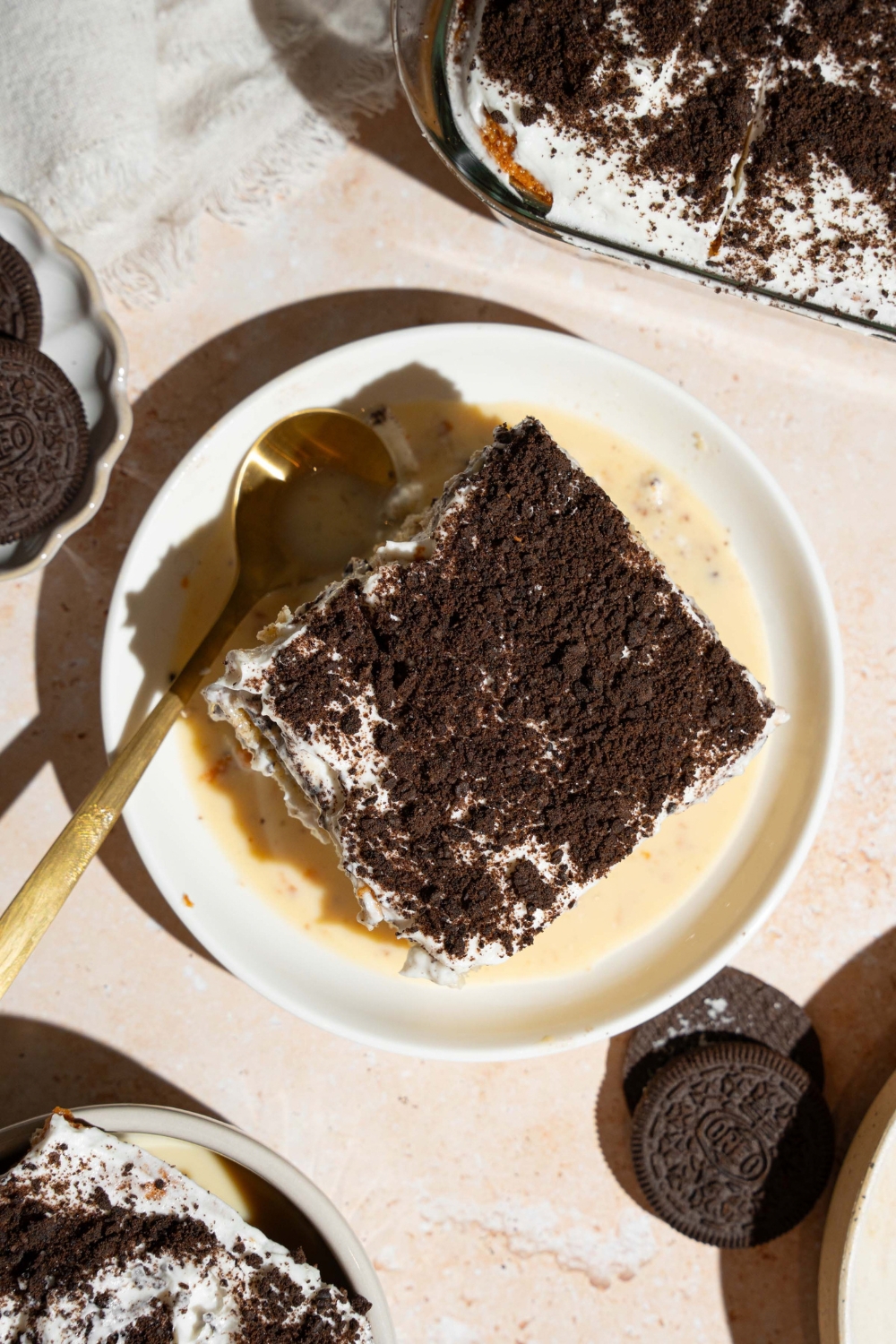A white plate with a slice of Oreo tres leches cake on top of tres leches soak. There is a spoon on the plate. The plate is on a tan counter with Oreos.